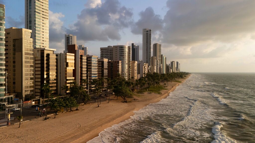 Foto aérea do DJI Mini 4 Pro da Praia de Boa Viagem, Recife, ao nascer do sol (06h). A imagem destaca a orla com prédios e palmeiras, capturada com filtro CPL e ND16 para cores vibrantes e reflexos reduzidos.