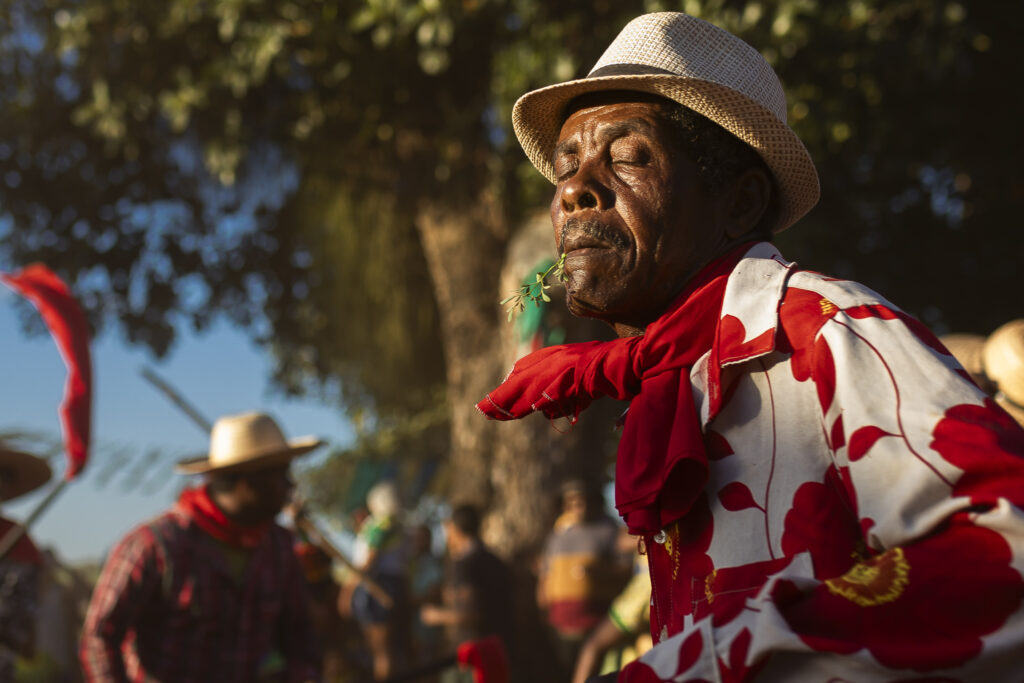 Fotografia de um brincante de Maracatu Rural, em close, com uma camisa vermelha e branca, e um chapéu de palha, com os olhos fechados em um momento de emoção durante a celebração do Cambinda Brasileira.