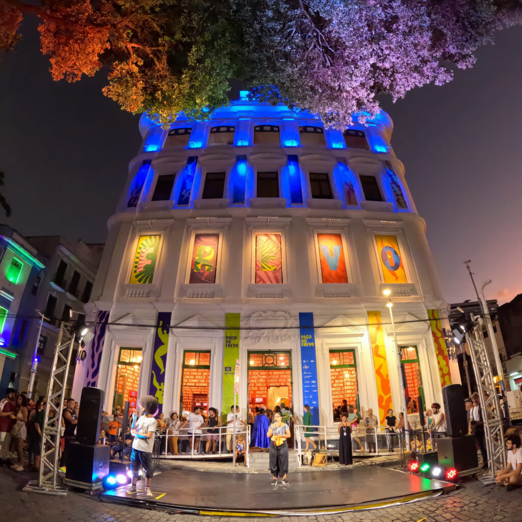 Fotografia em modo amplo com GoPro: vista noturna do Paço do Frevo, em Recife, com a fachada colorida por luzes de LED e um palco com público, capturando a arquitetura e a atmosfera do evento cultural.