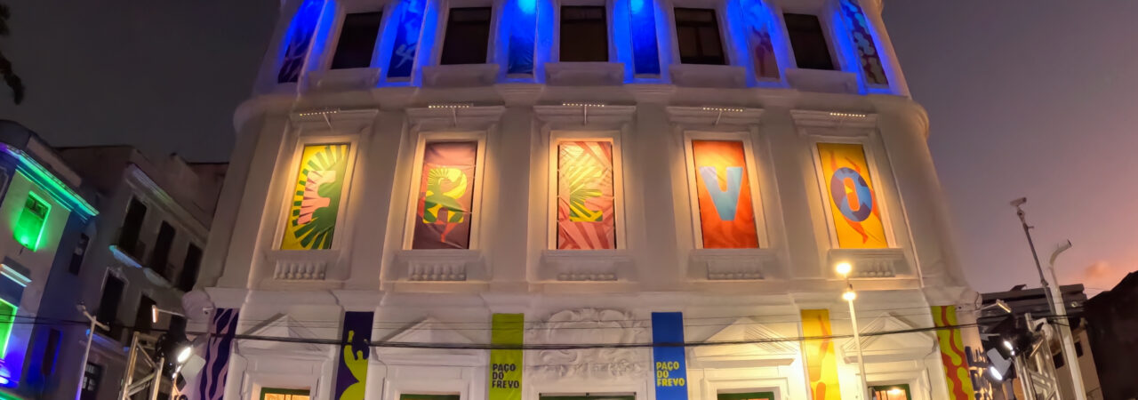 Fotografia em modo amplo com GoPro: vista noturna do Paço do Frevo, em Recife, com a fachada colorida por luzes de LED e um palco com público, capturando a arquitetura e a atmosfera do evento cultural.
