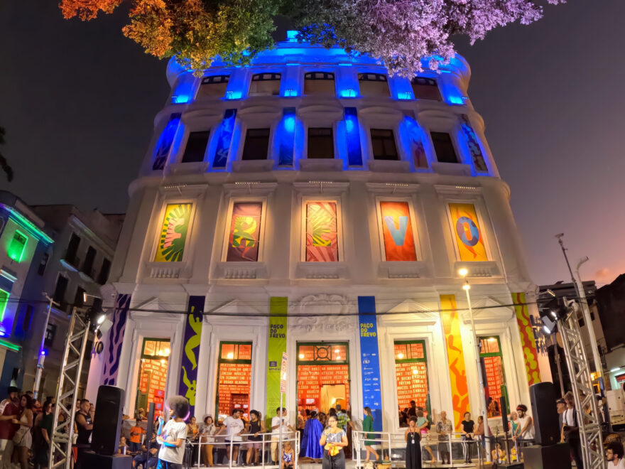 Fotografia em modo amplo com GoPro: vista noturna do Paço do Frevo, em Recife, com a fachada colorida por luzes de LED e um palco com público, capturando a arquitetura e a atmosfera do evento cultural.