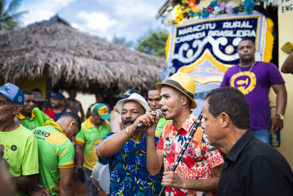 Foto documental do Maracatu Cambinda Brasileira, mostrando o Mestre do Apito Anderson Miguel e o Contra-Mestre cantando com o microfone, em meio aos brincantes da manifestação cultural.