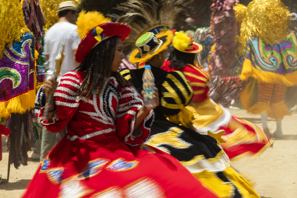Fotografia de manifestação cultural que utiliza o desfoque de movimento (motion blur) para capturar três dançarinas. A imagem destaca as cores vibrantes das fantasias (vermelho e amarelo) e o movimento circular das saias, transmitindo o ritmo e o dinamismo da dança popular.