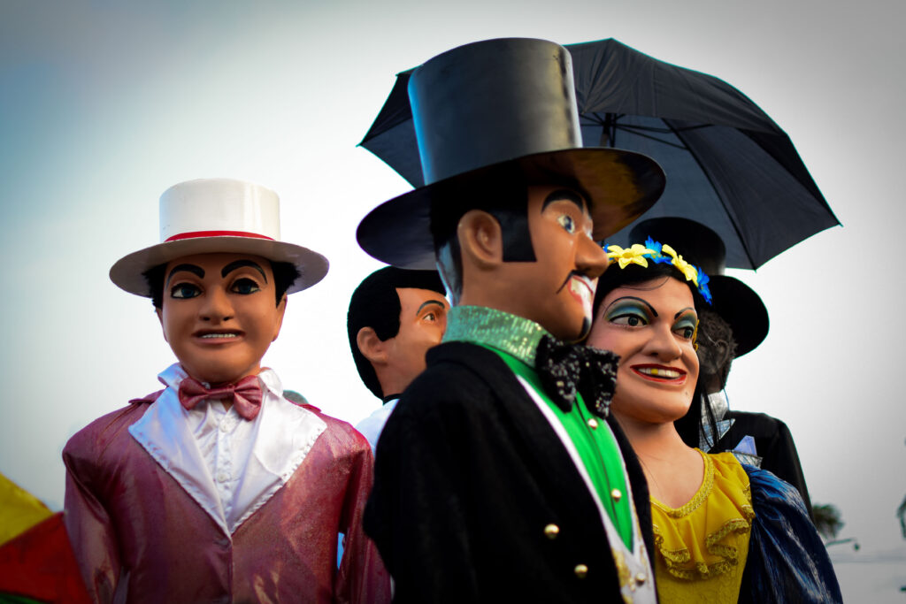 Close-up dos bonecos gigantes de Olinda, mostrando o casal de bonecos (o homem de cartola preta e terno preto com gravata borboleta verde, e a mulher de vestido amarelo e flores azuis) ao lado de outros bonecos. A imagem destaca as cores e a expressão dos personagens tradicionais do Carnaval de Pernambuco. Foto tirada com uma lente 35 mm e sensor APS-C, equivalente a lente 50 mm.
